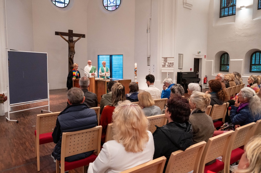 Gottesdienst in einer modernen Kirche mit Gemeinde in den Stuhlreihen und drei Personen in liturgischer Kleidung am Altar.