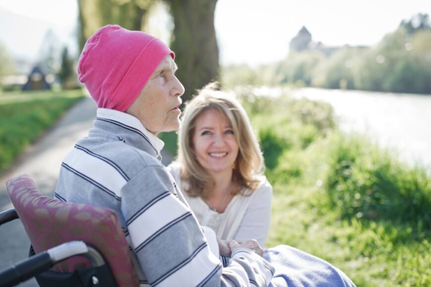 Eine ältere Frau mit pinker Mütze sitzt im Rollstuhl an einem Flussufer und blickt ins Profil, während eine lächelnde jüngere Frau sanft hinter ihr hockt.