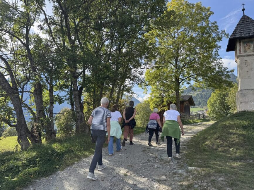 Eine Gruppe von Menschen wandert von hinten gesehen auf einem Kiesweg bergauf, vorbei an Bäumen und einem Bildstock.