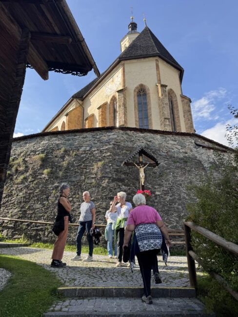 Eine Personengruppe steht auf einem gepflasterten Weg vor einer imposanten Steinmauer und einer Kirche mit Kruzifix.