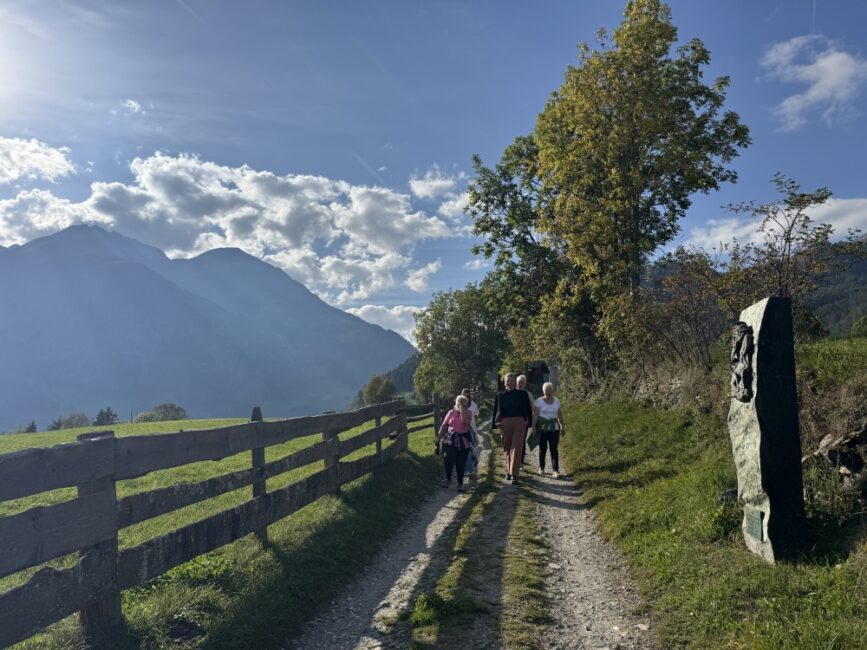 Auf dem Bild wandert eine Gruppe von mehreren Personen auf einem schmalen Schotterweg in einer sonnigen Berglandschaft. Der Weg verläuft leicht ansteigend und wird auf der linken Seite von einem niedrigen Holzzaun und einer saftig grünen Wiese begrenzt. Rechts des Weges steht ein hoher, aufrechter Gedenkstein mit eingemeißelten Figuren sowie ein größerer Baum mit teilweise herbstlich verfärbten Blättern. Im Hintergrund erhebt sich ein imposantes, massives Bergmassiv unter einem blauen Himmel mit vereinzelten, hellen Wolken.
