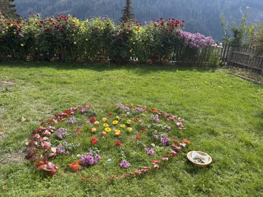 Auf einer grünen Wiese gelegtes Natur-Mandala aus bunten Herbstblättern, Blumen und Äpfeln vor einem üppigen Blumenbeet.