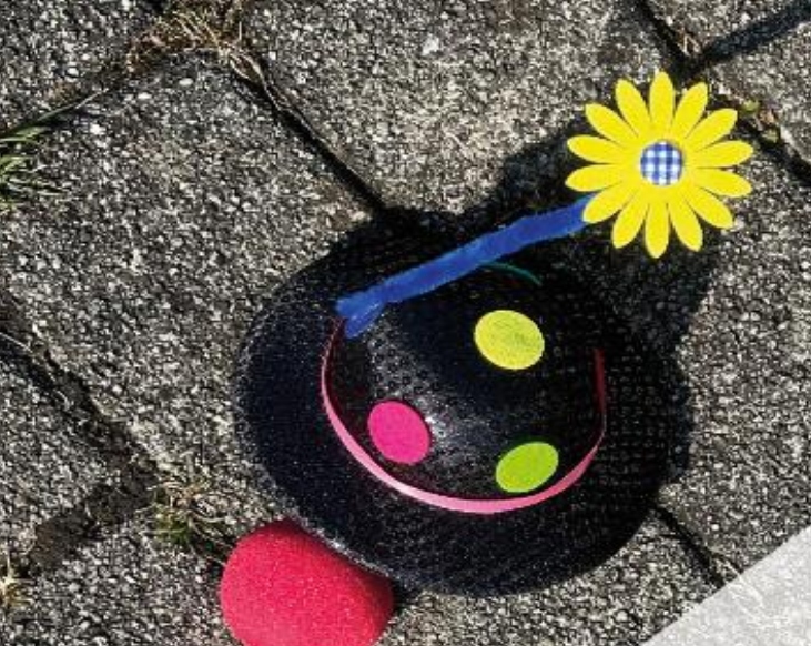 Ein kleiner schwarzer Clownshut mit bunten Punkten, einer gelben Blume am Stiel und einer roten Clowns Nase, die auf einem asphaltierten Gehweg liegen.