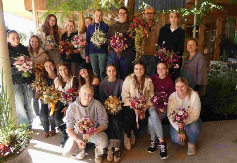 Eine Gruppe junger Schüler*innen steht und sitzt im Freien vor dem Hospizhaus und präsentiert stolz ihre selbst gebundenen, bunt gestalteten Blumensträuße.